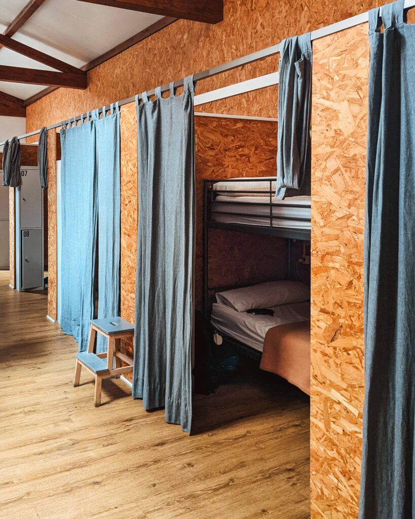 Dorm room with alcoves of bunk beds that have floor-length blue privacy curtains at Albergue a Conserveira