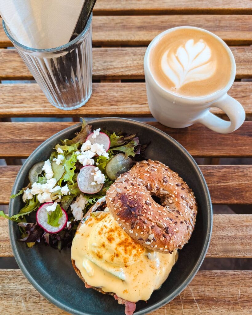 Top down view of a latte in a white cup, blue plate with a bagel, poached egg, hollandaise sauce, bacon, and salad with radish and cheese. A clear cup holds napkins and cutlery.