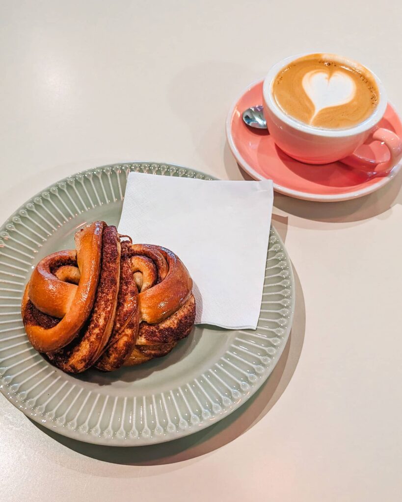 Latte in a pink cup and saucer, and a Swedish cinnamon roll on a light green plate with a paper napkin.