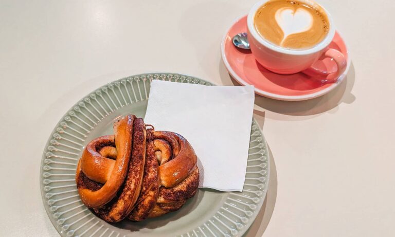 Latte in a pink cup and saucer, and a Swedish cinnamon roll on a light green plate with a paper napkin.