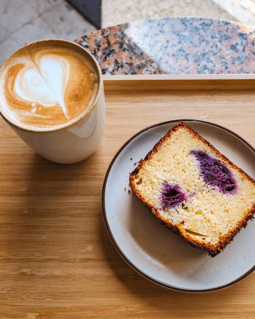 Top down view of a latte and a lemon and berry pound cake slice served on a wooden tray on top of an orange and gray marble table.
