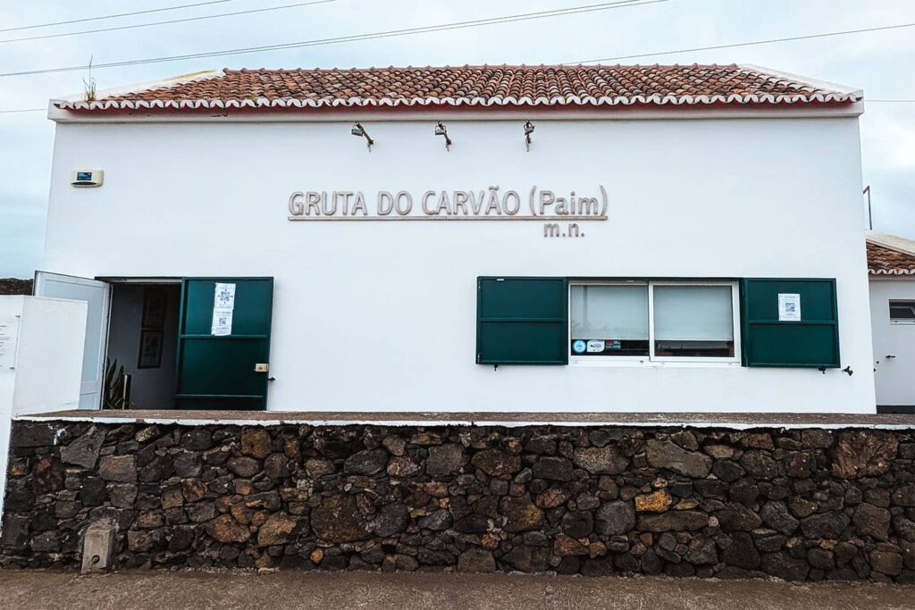 Front of Gruta do Carvao office. A white building with green window shutters, a green door, and an orange tile roof with a brick wall in front.