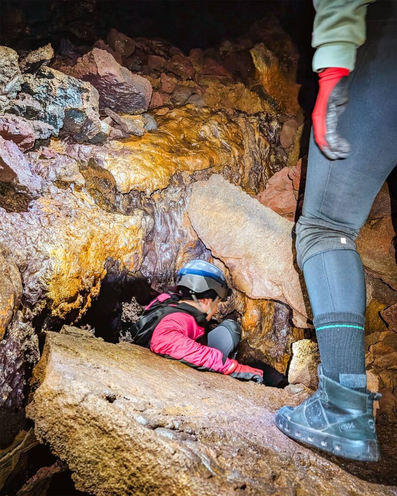 Girl in a pink jacket, gloves, and helmet descends into a small rock opening as someone stands to the side in Gruta do Carvao.
