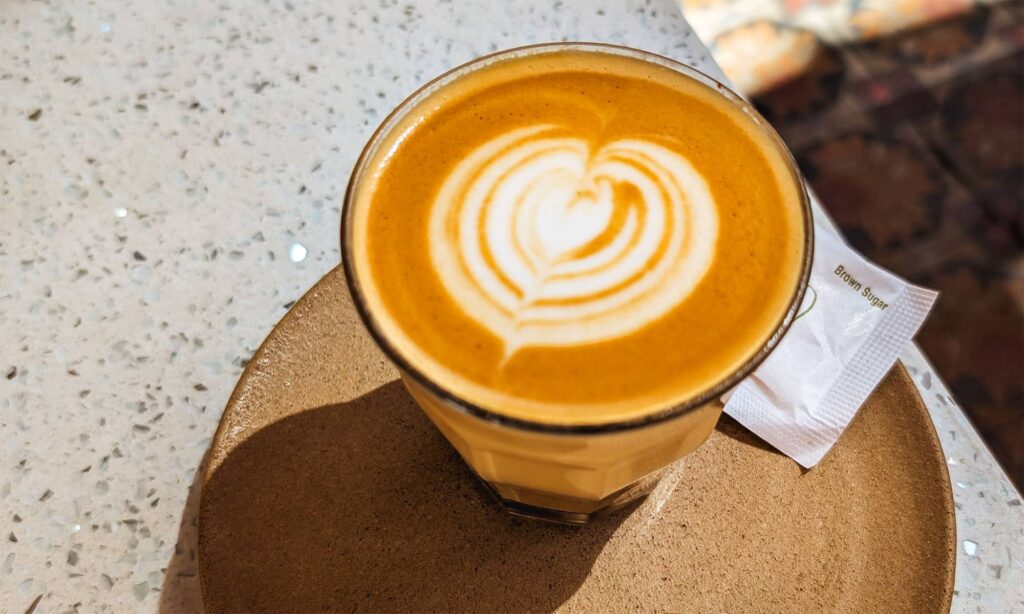 Latte art in a glass cup and a packet of brown sugar on a brown ceramic saucer at Oso Cafe in Arequipa, Peru