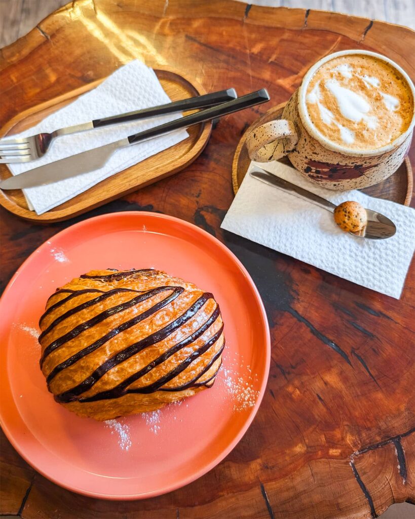 Top down view of a croissant with chocolate stripes on a pink plate and a ceramic mug with latte art on a wood table with utensils